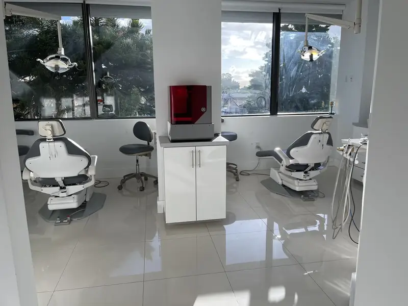 Bright operatory with two dental chairs and the in-house 3D printer under a translucent red cover